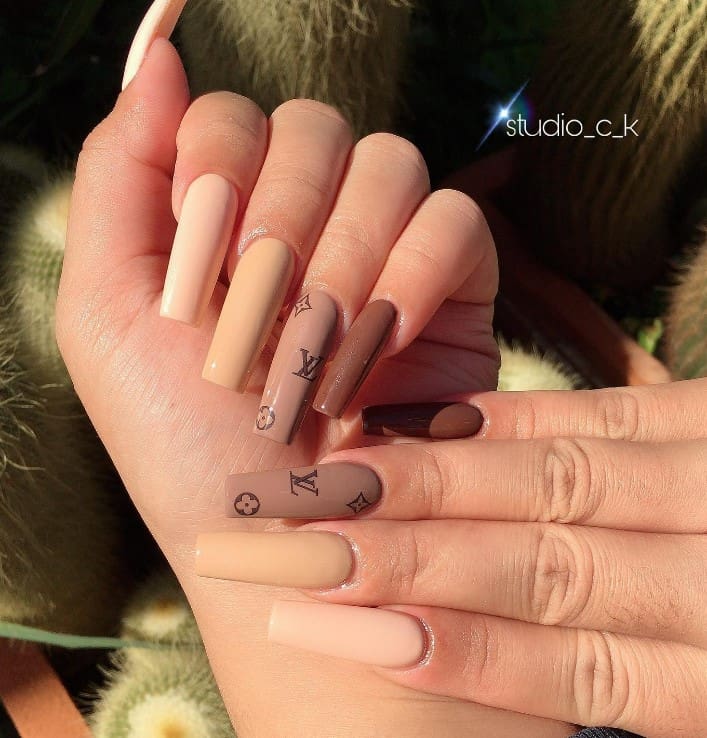 A closeup of a woman's fingernails with a different shades of brown nail polish that has  luxury brand logos to one nail