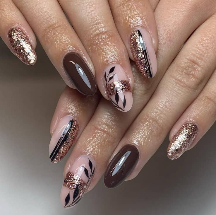 A closeup of a woman's fingernails with a deep brown and nude nail polish that has rose gold shimmer and black leaf nail art 