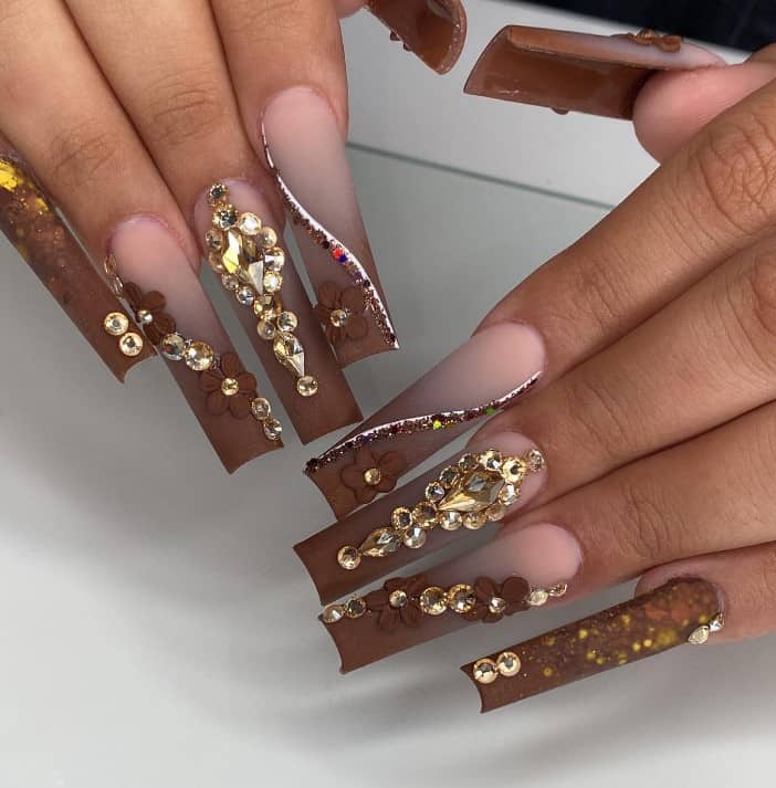 A closeup of a woman's fingernails with a nude ombre nail polish that has brown tips, rhinestones, flowers, and 3D art