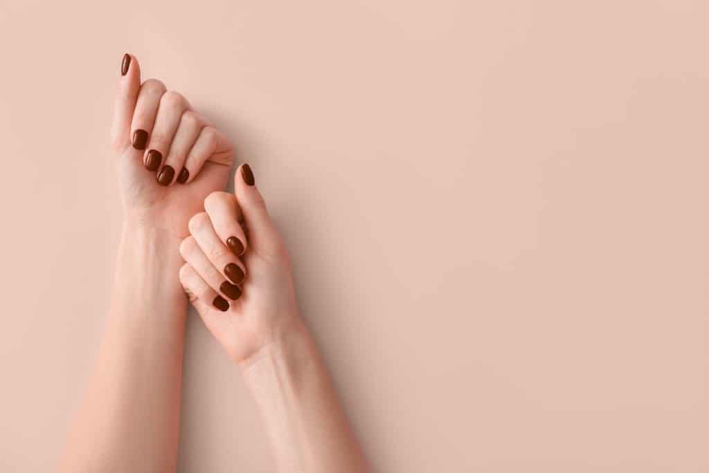 A woman's fingernails with a dark shade of brown nail polish isolated on a peach background