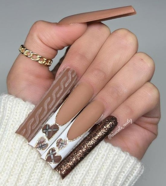 A woman's fingernails with a nude brown nail polish and brown glitter polish to one nail that has knitted sweater patterns on another and white French tips with brown plaid hearts and outlined with gold glitter