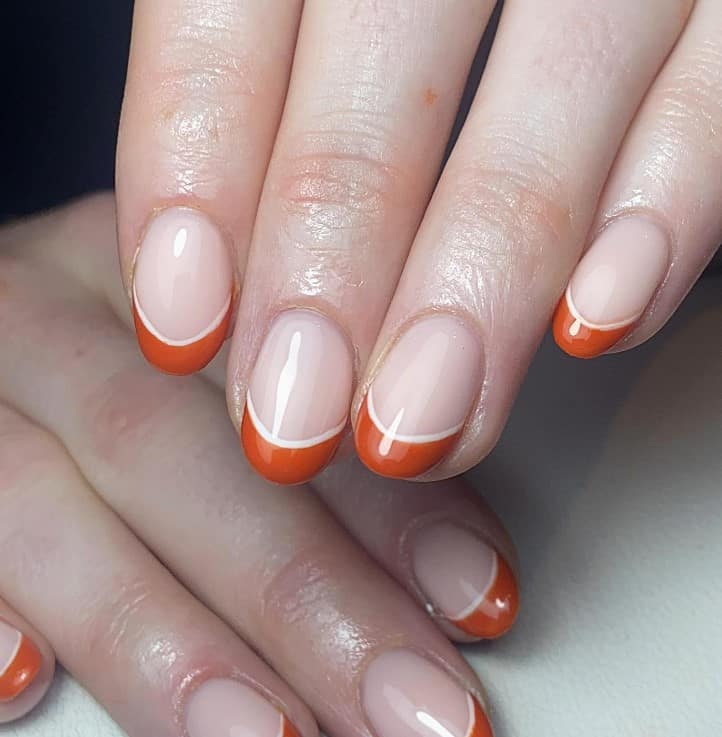 A closeup of a woman's round-shaped fingernails with a glossy nude nail polish base that has burnt orange French tips with crisp white lines outlining the bottom of the tips