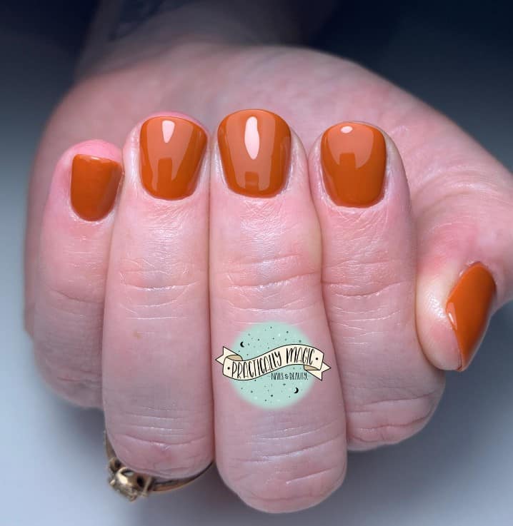 A woman's short fingernails with a glossy burnt orange nail polish base 