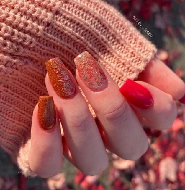 A closeup of a woman's short coffin fingernails with glittery burnt orange nails and a bold red accent nail that has bits of foil on orange nails