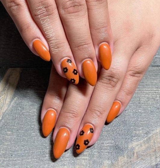 A closeup of a woman's almond-shaped fingernails with a burnt orange nail polish base that has stamped black florals on select nails 