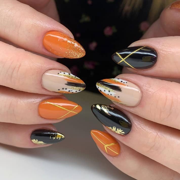 A closeup of a woman's stiletto shaped fingernails with a combination of burnt orange, cream, and black nail polish that has smeared tips, gold leaves, fine glitter, and gold crisscross patterns