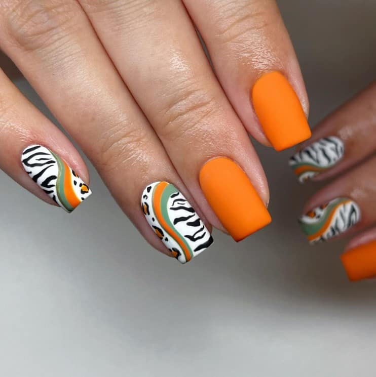 A woman's fingernails with a combination of a matte burnt orange and white nail polish base that has leopard and zebra prints, divided by green and orange swirls