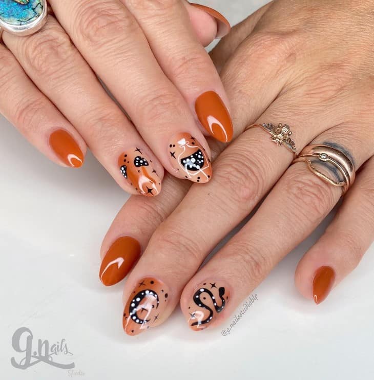 A woman's fingernails with a solid burnt orange nail polish that has black-and-white funky designs like twinkling stars, potion bottles, crescent moons, and snakes nail designs