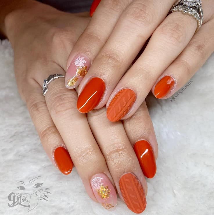 A woman's oval shaped fingernails with a burnt orange nails that has a knit texture in matte orange, delicate fall leaves in white and gold on natural-colored nails