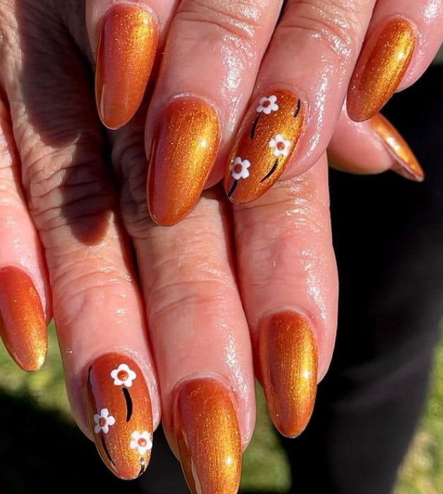 A closeup of a woman's oval fingernails with burnt orange hue nail polish that has stemmed white flowers on select nails