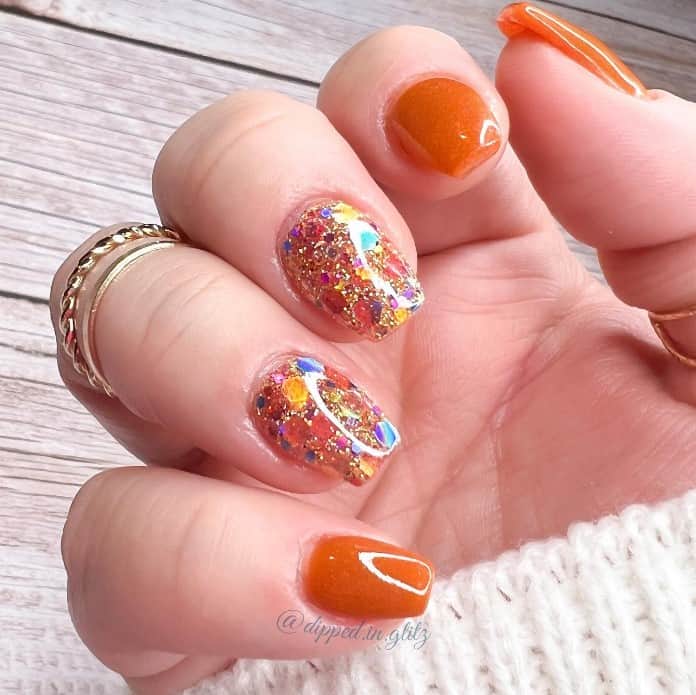 A closeup of a woman's fingernails with a a glossy burnt orange nails and chunky glitter on clear polish on select nails