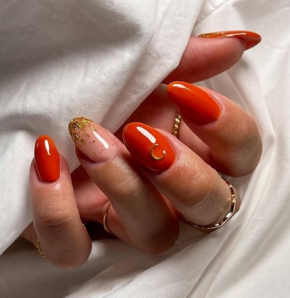 A woman's almond fingernails with a combination of peach and burnt orange nail polish that has gold flecks on the peach-colored nail and gold crescent moon stud on the middle nail