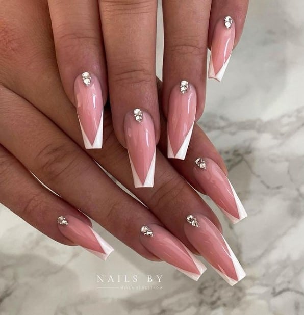 A closeup of a woman's fingernails with a nude nail polish base that has white chevron French tips and glittery rhinestone accents