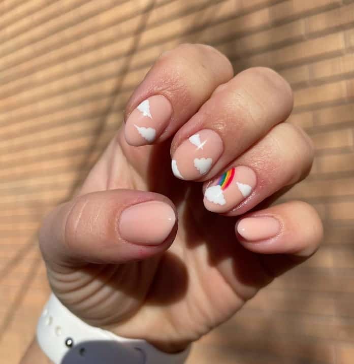 A woman's fingernails with a creamy ɴuᴅᴇ nail polish base that has clouds and rainbows nail designs on select nails 