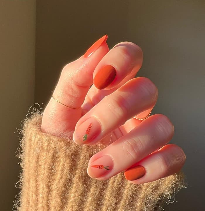 A woman's fingernails with deep orange and nude polish that has carrot nail art on select nails