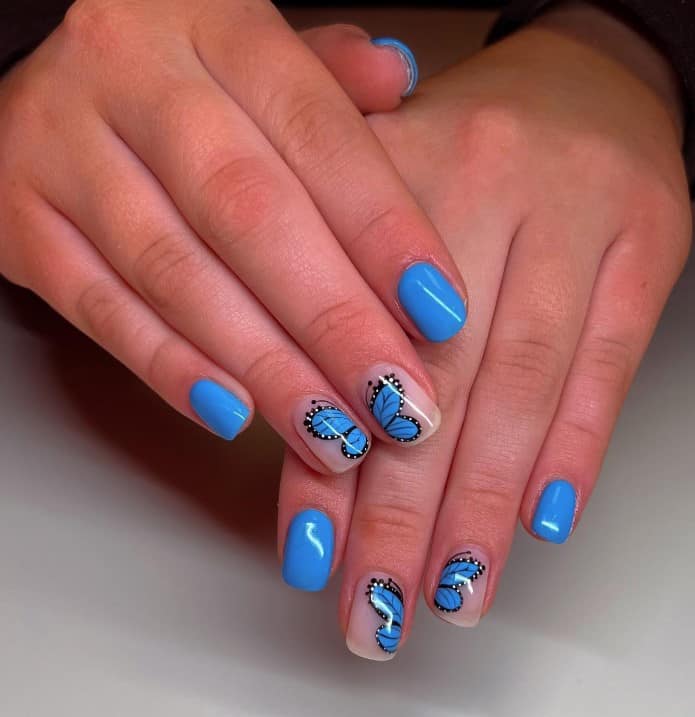 A woman's fingernails with a combination of electric blue and nude nail polish that has blue butterfly on select nails 