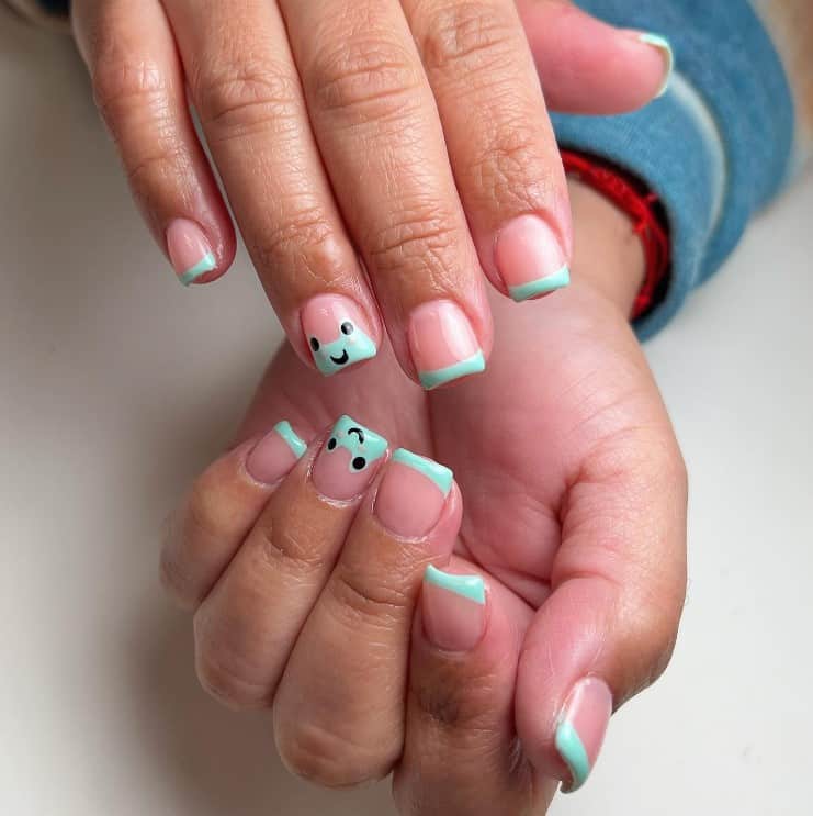 A woman's fingernails with a nude nail polish base that has blue French tips with fun froggy nail art