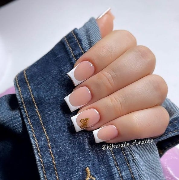 A woman's fingernails with a beautiful nude nail polish that has white tips and a gold butterfly on select nail