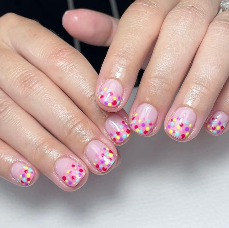 A closeup of a woman's fingernails with a nude nail polish that has multicolored dots on nail tips 