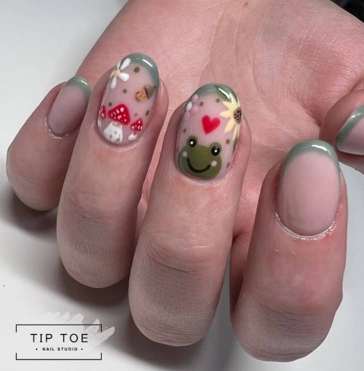 A closeup of a woman's fingernails with a nude nail polish that has gray-green tips, frogs, mushrooms, and other nature-inspired elements nail designs on select nails