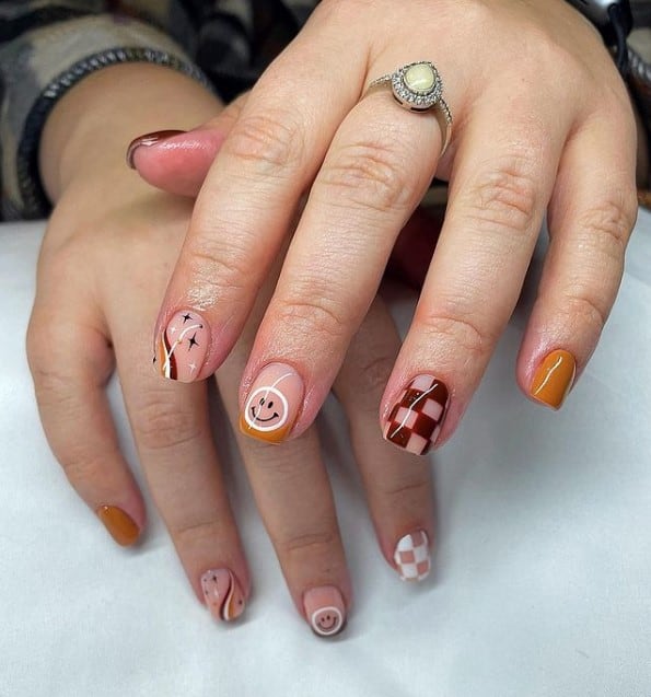 A closeup of a woman's fingernails with a nude nail polish base that has swirls, checkered patterns, and smiley face nail designs