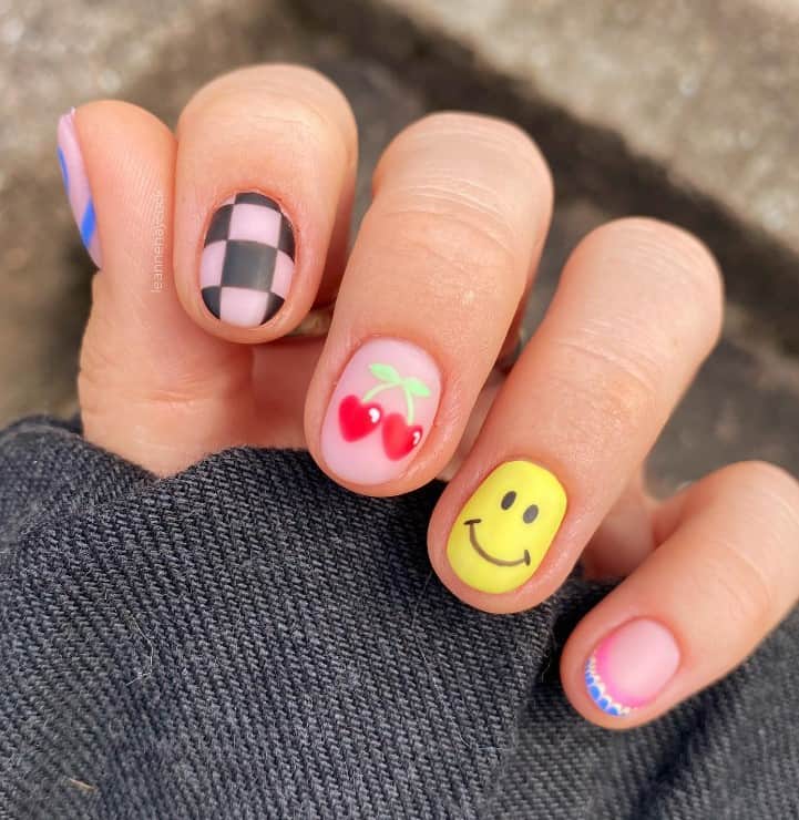 A woman's fingernails with a nude nail polish base that has cherries, smiley faces, or checkered nail designs 