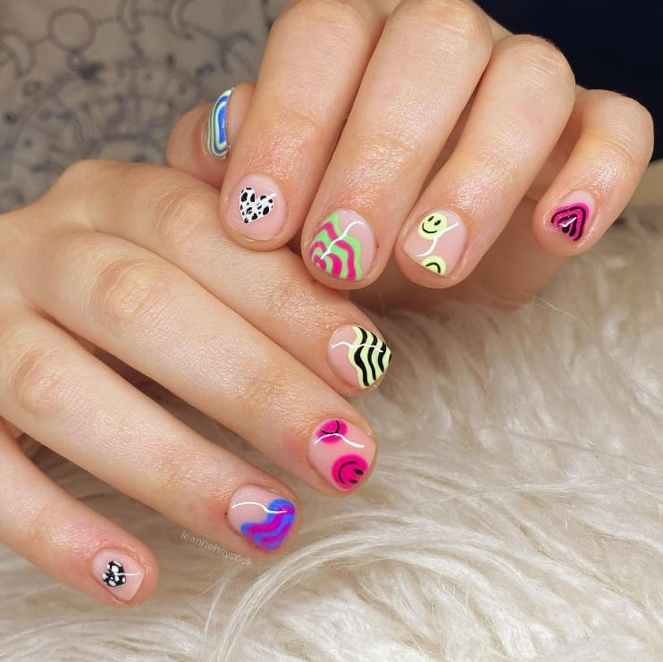 A woman's fingernails with a nude nail polish base that has bright swirls, smiley faces, and patterned hearts nail designs