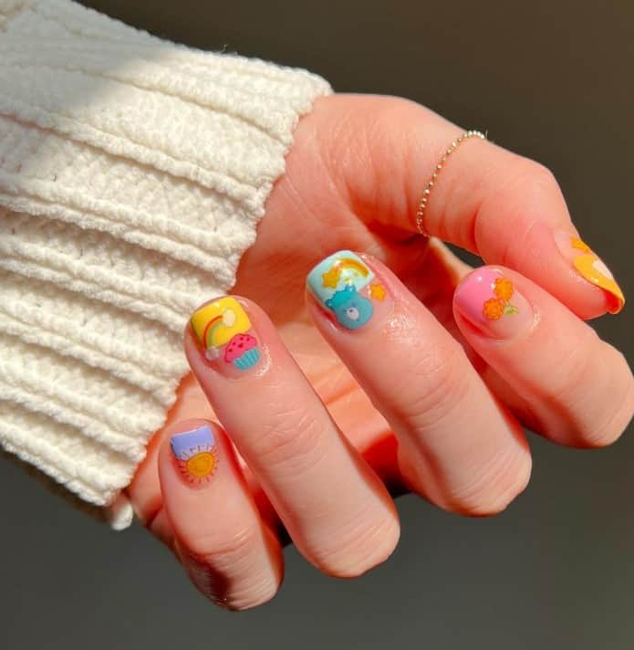 A closeup of a woman's fingernails with a nude nail polish base that has Care Bear, cupcakes, flowers, rainbows, and stars nail designs