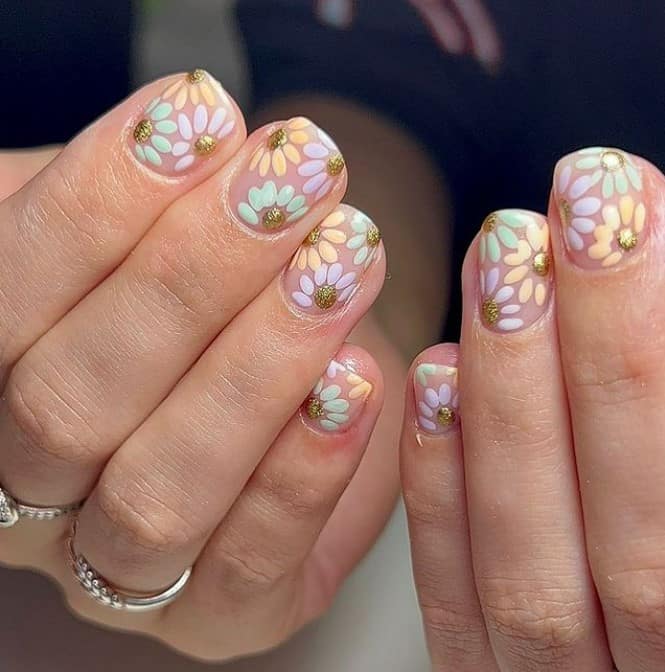 A closeup of a woman's fingernails with a nude nail polish that has pastel-hued daisies and glittery gold polish nail designs