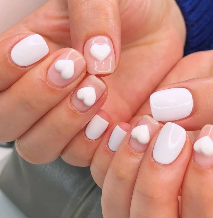A closeup of a woman's fingernails with a combination of nude and white nail polish that has 3D hearts