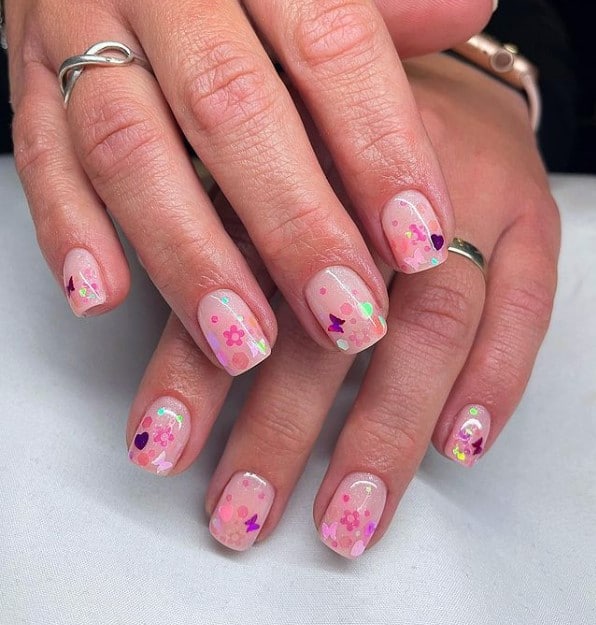 A closeup of a woman's fingernails with a pale pink nail polish that has flower, butterfly, and heart stickers nail designs