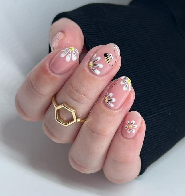 A closeup of a woman's short oval fingernails with a beautiful nude nail polish that has bees and flowers nail designs