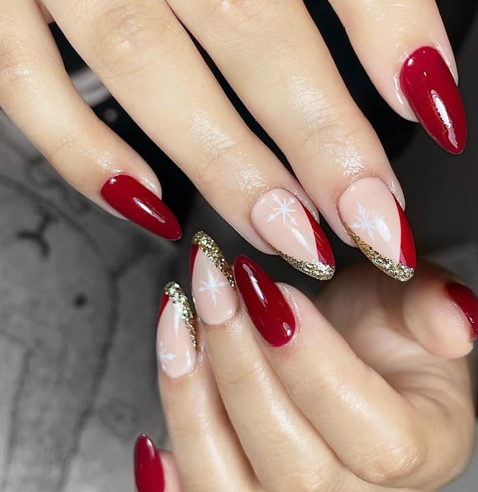 A closeup of a woman's fingernails with a combination of blood red and nude nail polish that has white snowflake and half gold glittery French tips on select nails 