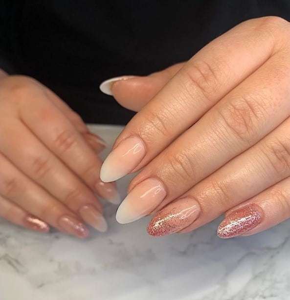 A closeup of a woman's fingernails featuring white and nude peach ombré nails that has rose gold glitter on select nails 