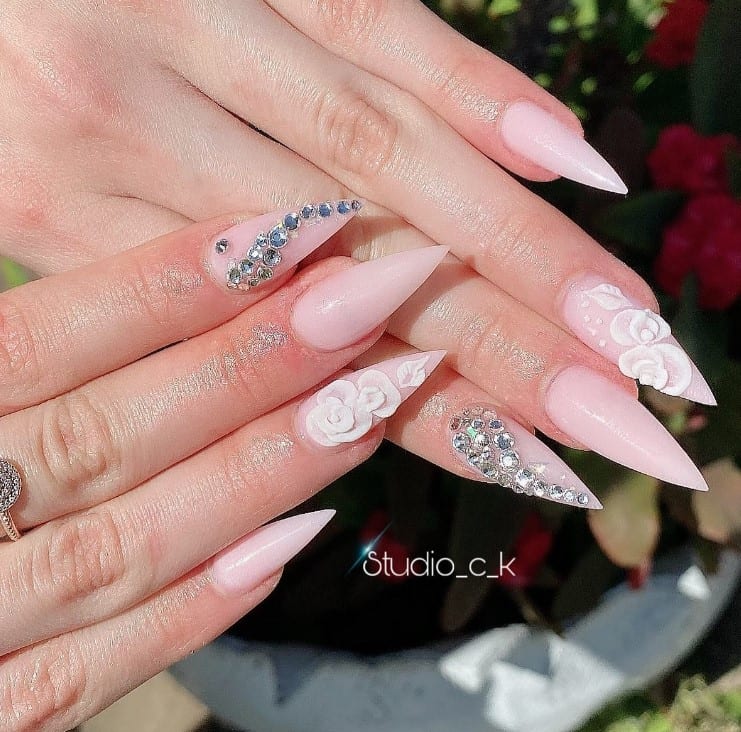A closeup of a woman's fingernails with pale pink base color that has 3D white roses and rhinestones on select nails 