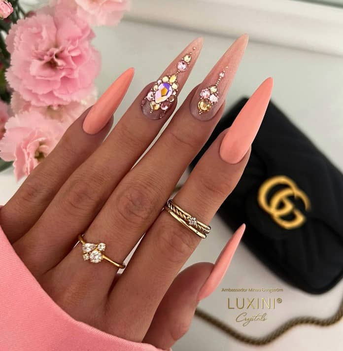 A closeup of a woman's stiletto fingernails with peach nail polish that has diamonds and gems on the middle and ring fingernails