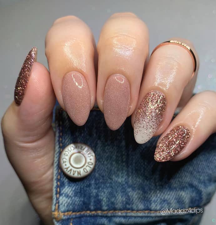 A closeup of a woman's fingernails with combination of nude shades and a white accent nail that has rose gold glitter on accent nail