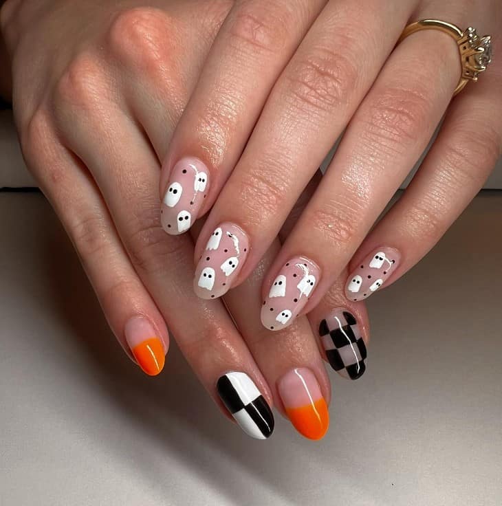 A closeup of a woman's fingernails with a combination of nude and checkered black-and-white patterns nail polish that has ghost designs and alternating blocks of bold orange nail designs