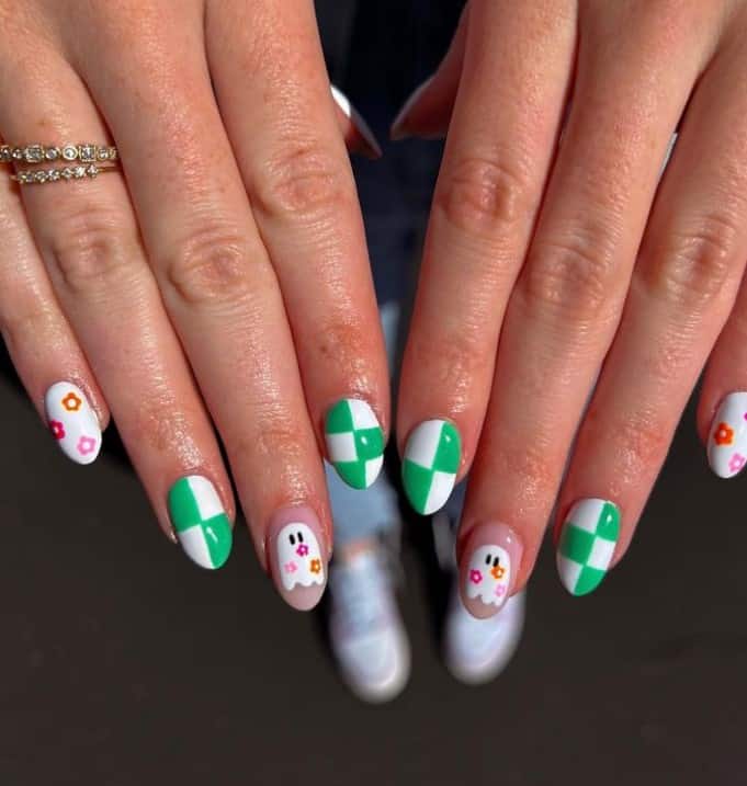 A closeup of a woman's fingernails with a white and peach nail polish base that has green and white checkered nails and white ghosts with flower accents