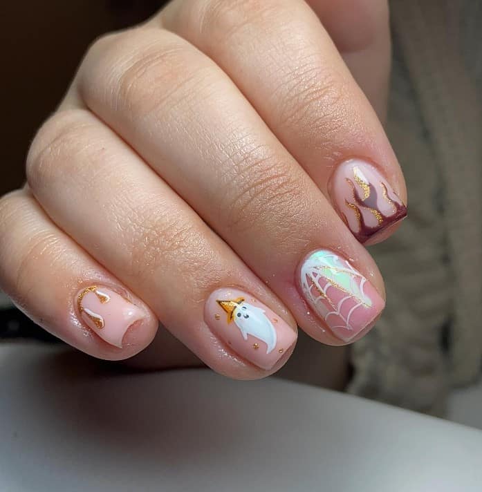 A closeup of a woman's fingernails with a light pink base that has sparkling glittery gold and white accents such as ghosts, spiderwebs, and eerie drippings nail designs