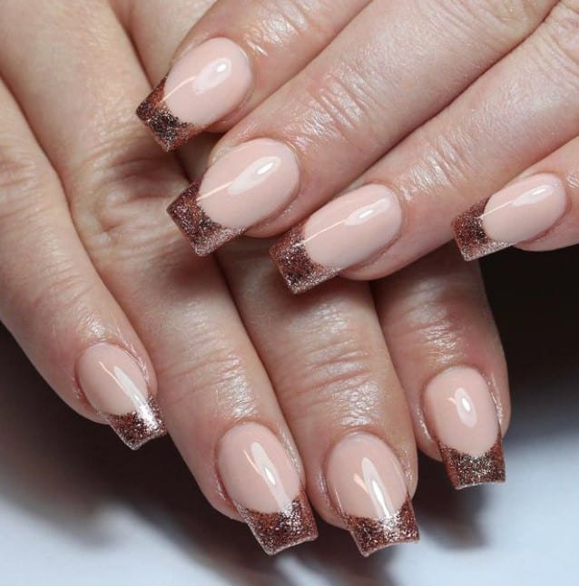 A closeup of a woman's fingernails with a glossy clear base that has darker shade of rose gold glitter for the French tips