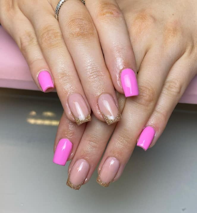 A closeup of a woman's fingernails with a combination of nude and bright pink nail polish base that has gold glitter French tips