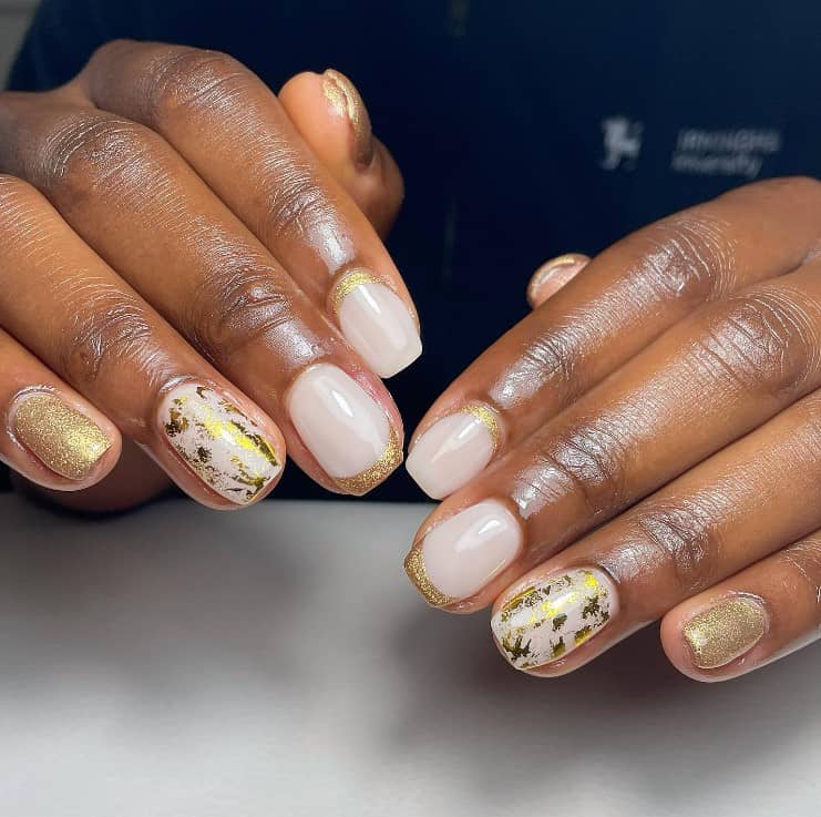 A closeup of a woman's fingernails with a combination of white and gold nail polish base that has gold flakes, gold French tips and gold cuticle cuffs