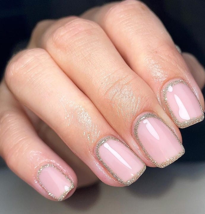 A closeup of a woman's fingernails with a glossy nude nail polish base that has gold glitter border