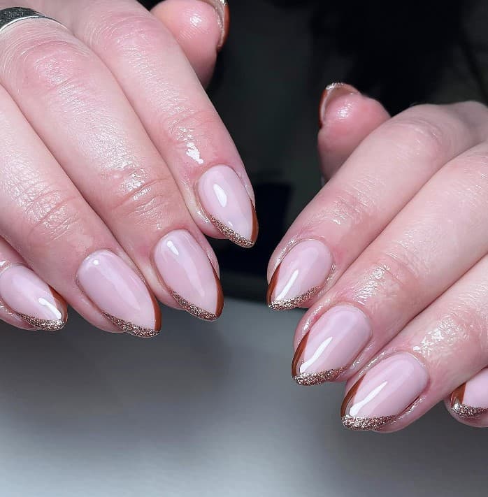 A closeup of a woman's fingernails with a glossy nude nail polish base that has V-shaped solid brown French tips topped with gold glitter