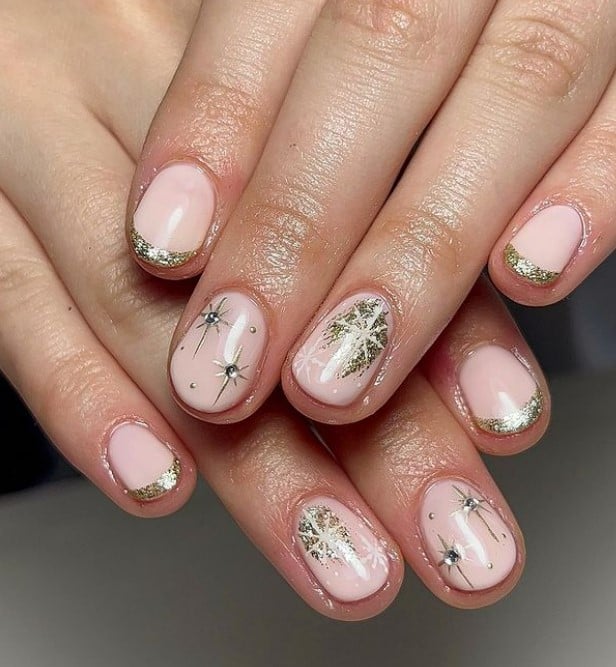 A closeup of a woman's fingernails with a nude nail polish base that has glittery gold French tips, intricate gold foil designs, and large gold stars with small gems