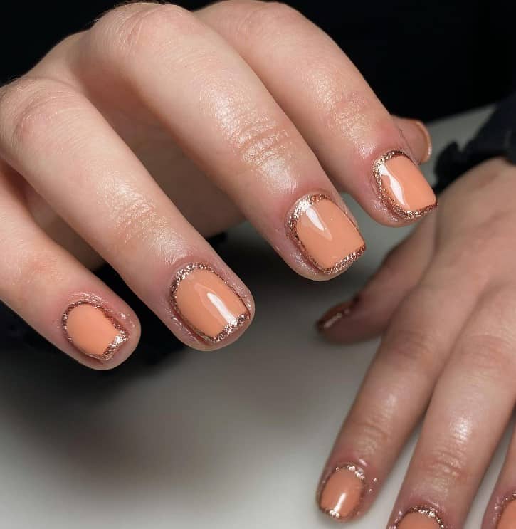 A closeup of a woman's fingernails with a peach nail polish base that has glittery rose gold frames