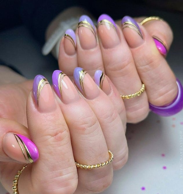 A closeup of a woman's fingernails with a nude nail polish base that has side French tips in different shades of purple with double outlines in gleaming gold
