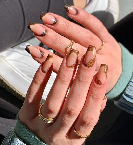 A woman's fingernails with a nude nail polish base that has glittery gold and coffee hues to paint thick waves across the tips and cuticles