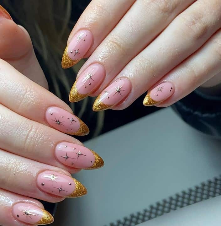 A closeup of a woman's almond fingernails with a nude nail polish base that has handpainted stars and tiny gems and gold French tips